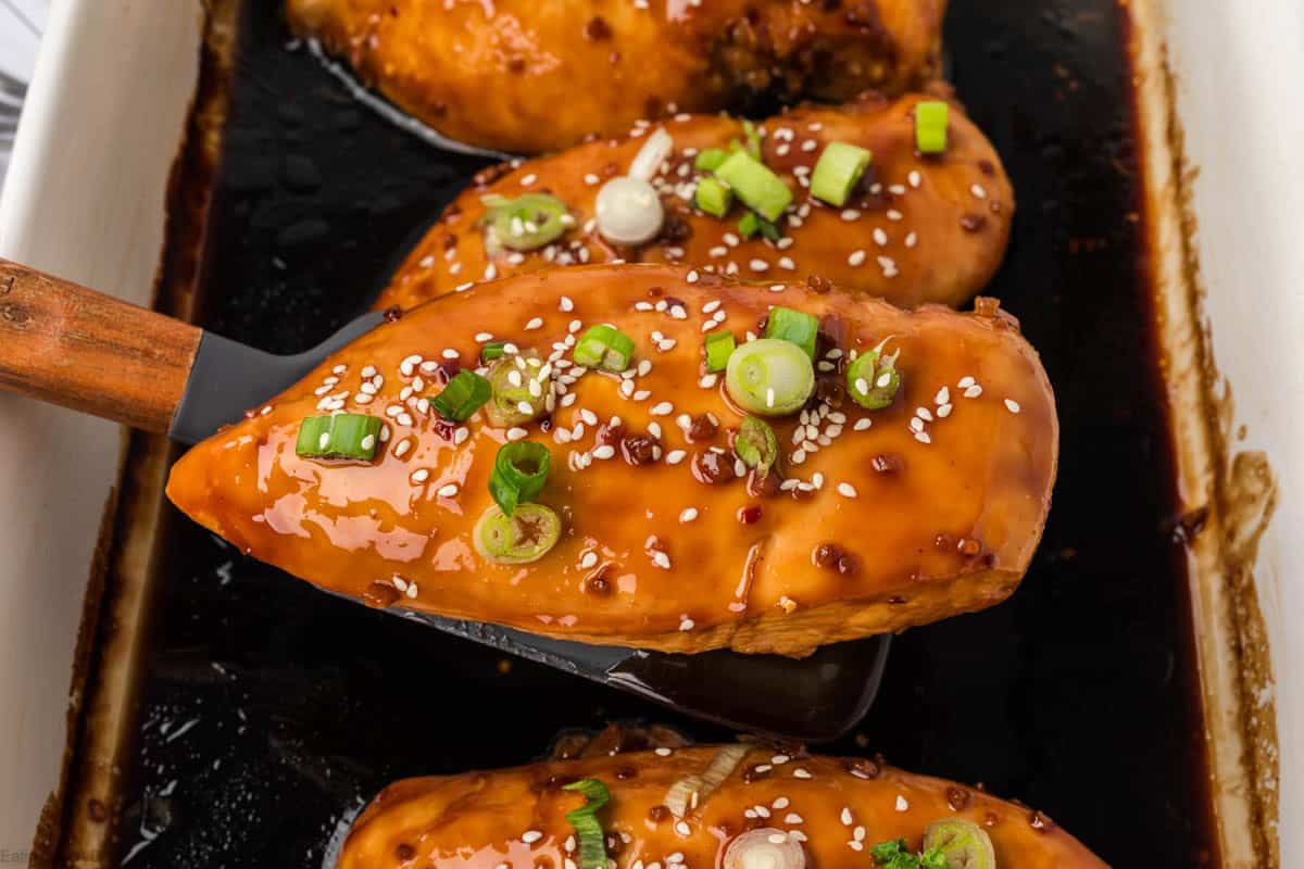 A spatula lifts a glazed chicken breast topped with sesame seeds and sliced green onions from a baking dish with more baked teriyaki chicken recipe pieces.