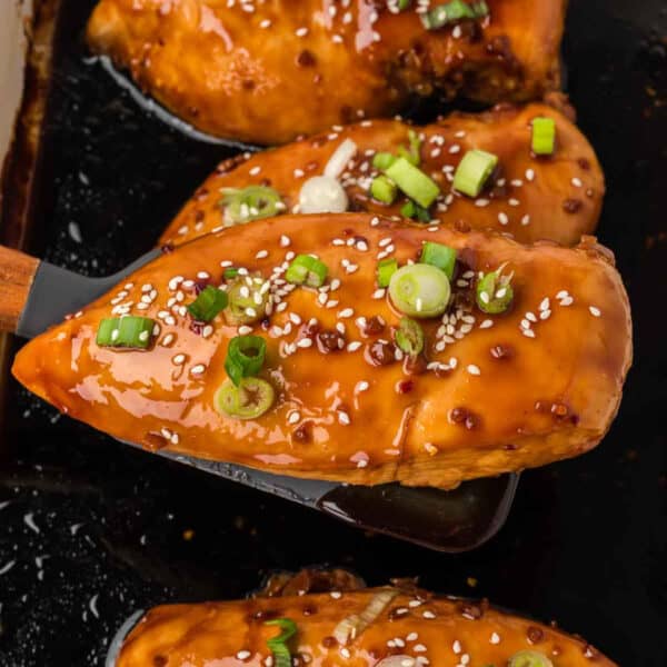 A close-up of glazed baked teriyaki chicken breasts topped with sesame seeds and sliced green onions, with one piece being lifted by a spatula from a dark baking dish.