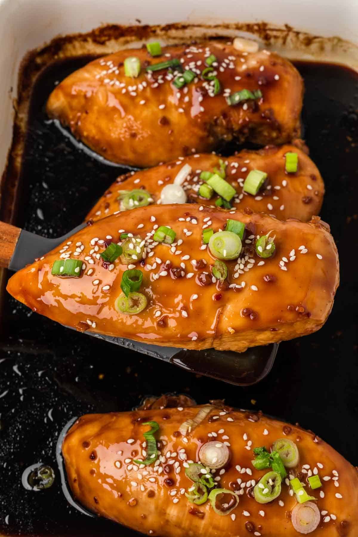 Three glazed baked chicken breasts in a dark teriyaki sauce, garnished with sliced green onions and sesame seeds, with one piece being lifted on a spatula from a baking dish—a delicious baked teriyaki chicken recipe.