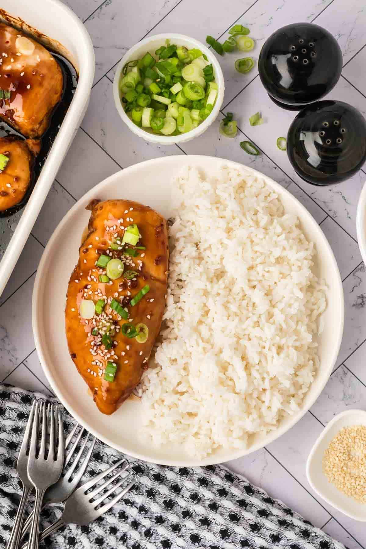 A plate of white rice and glazed chicken breast from a baked teriyaki chicken recipe, topped with sesame seeds and chopped green onions, is served beside a baking dish, salt and pepper shakers, and forks on a patterned napkin.