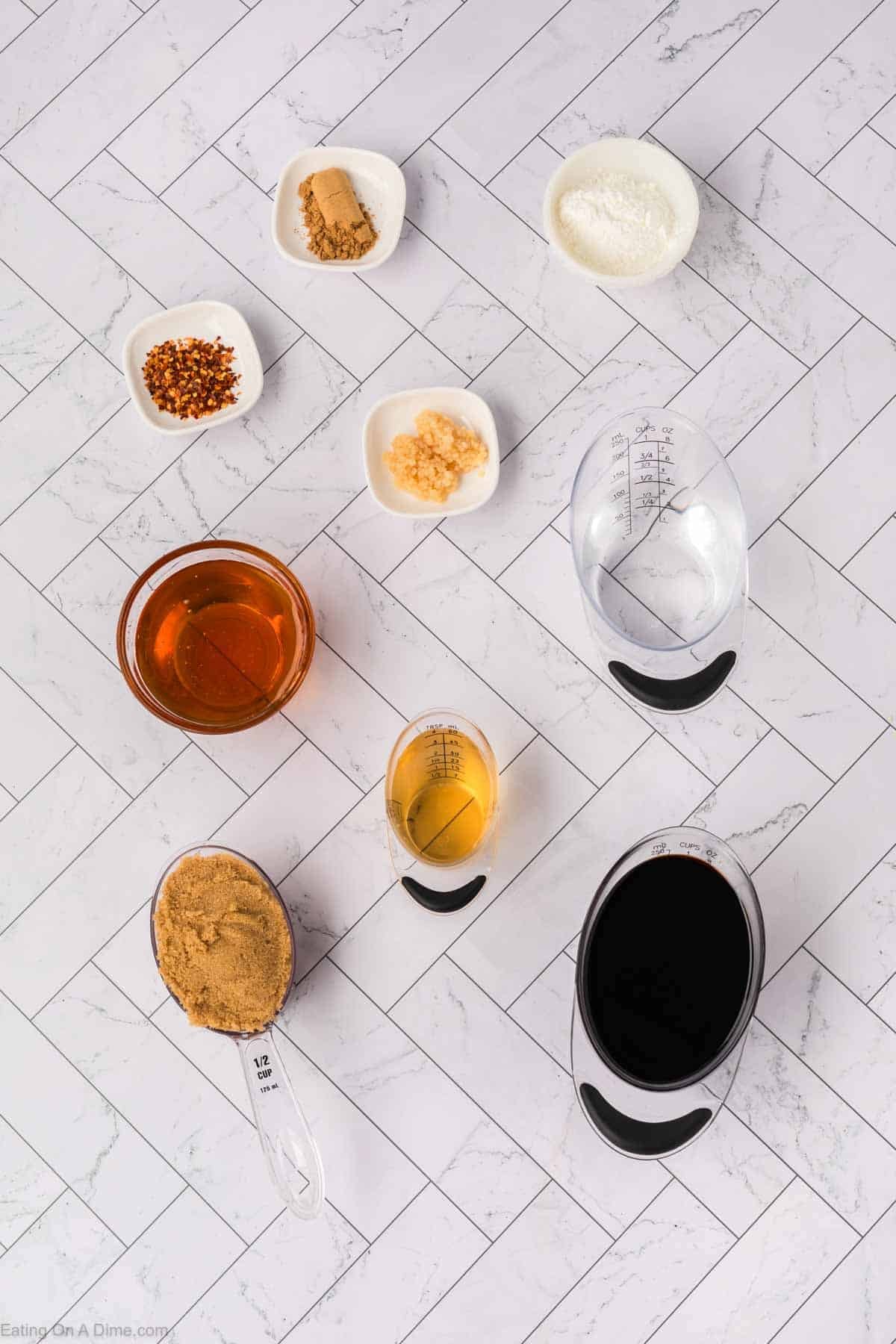 Top-down view of measured ingredients for a baked teriyaki chicken recipe in bowls and cups on a white tiled surface, including brown sugar, minced garlic, red pepper flakes, soy sauce, honey, cornstarch, apple cider vinegar, and ginger.