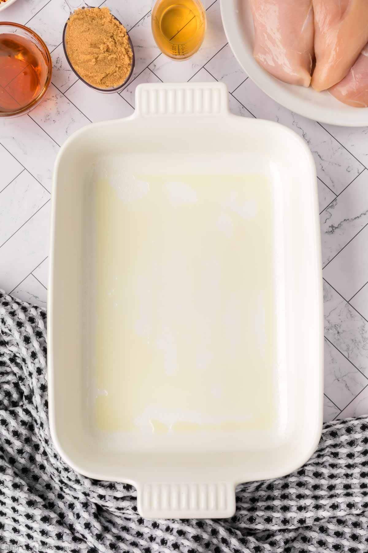 A greased white rectangular baking dish on a patterned surface, surrounded by raw chicken breasts, brown sugar, a small glass of honey, and a drink—perfect for preparing your favorite baked teriyaki chicken recipe—with a black and white cloth nearby.
