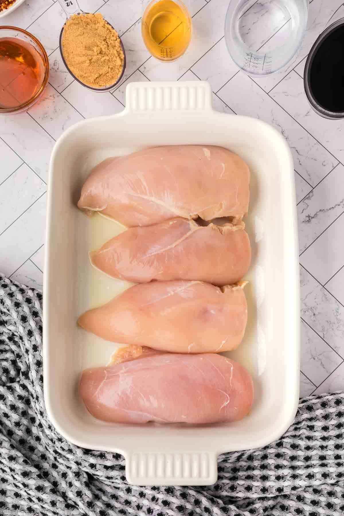 Four raw chicken breasts are arranged in a white baking dish, ready for this easy baked teriyaki chicken recipe. Surrounding the dish are small bowls of brown sugar, soy sauce, oil, and other marinade ingredients, with a black and white towel beside it.