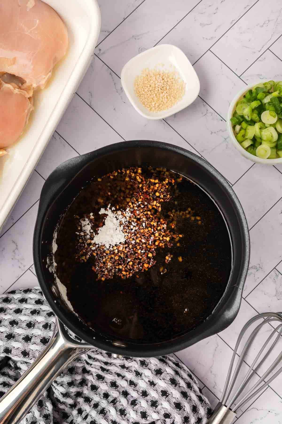 A saucepan filled with dark liquid, red pepper flakes, and seasonings sits on a tiled countertop—the start of a baked teriyaki chicken recipe. Nearby are green onions in a bowl, sesame seeds in a dish, raw chicken on a tray, and a whisk on a patterned towel.