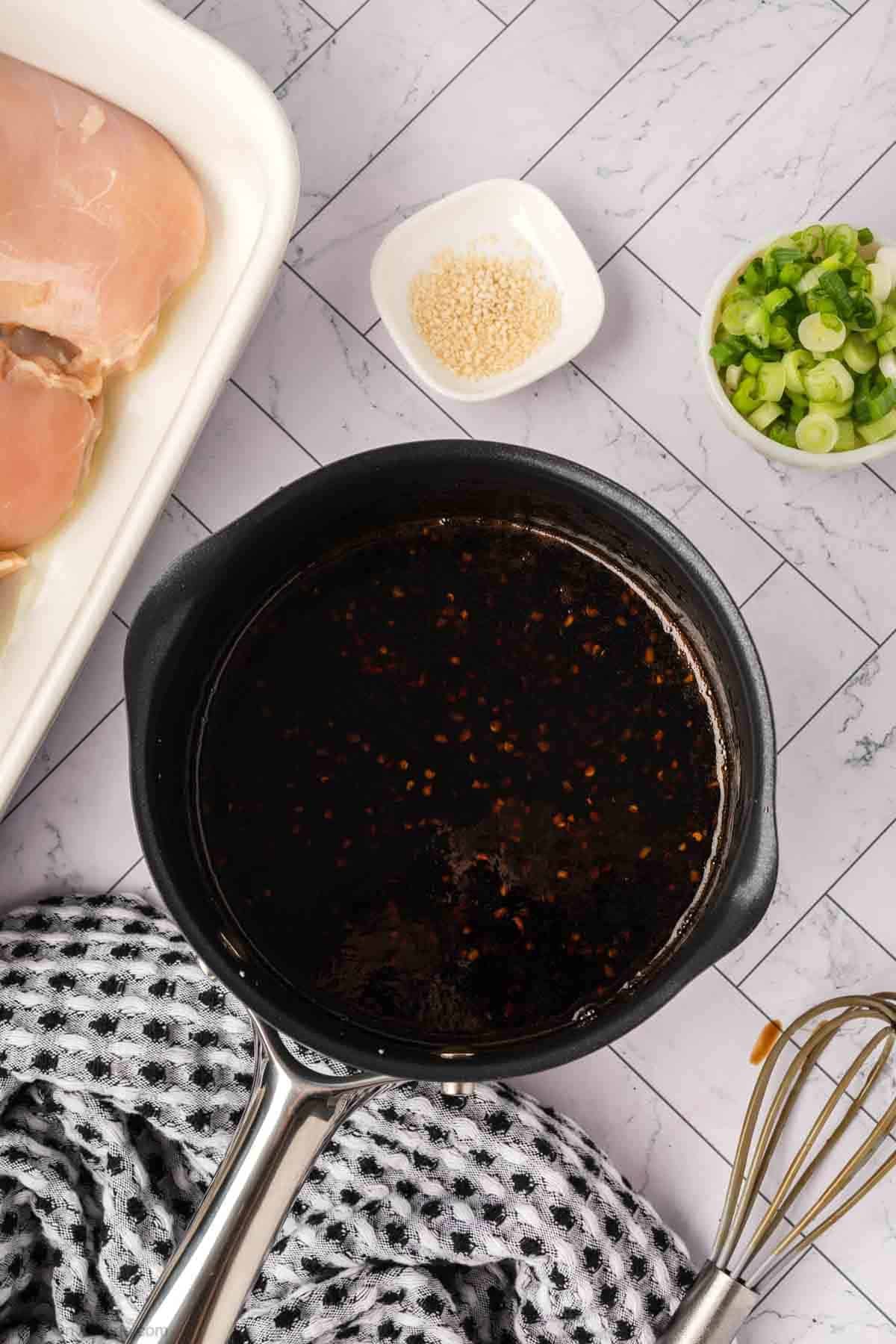 A saucepan filled with dark marinade sits on a tiled surface, surrounded by raw chicken ready for a baked teriyaki chicken recipe, chopped green onions, sesame seeds, a whisk, and a black-and-white towel.