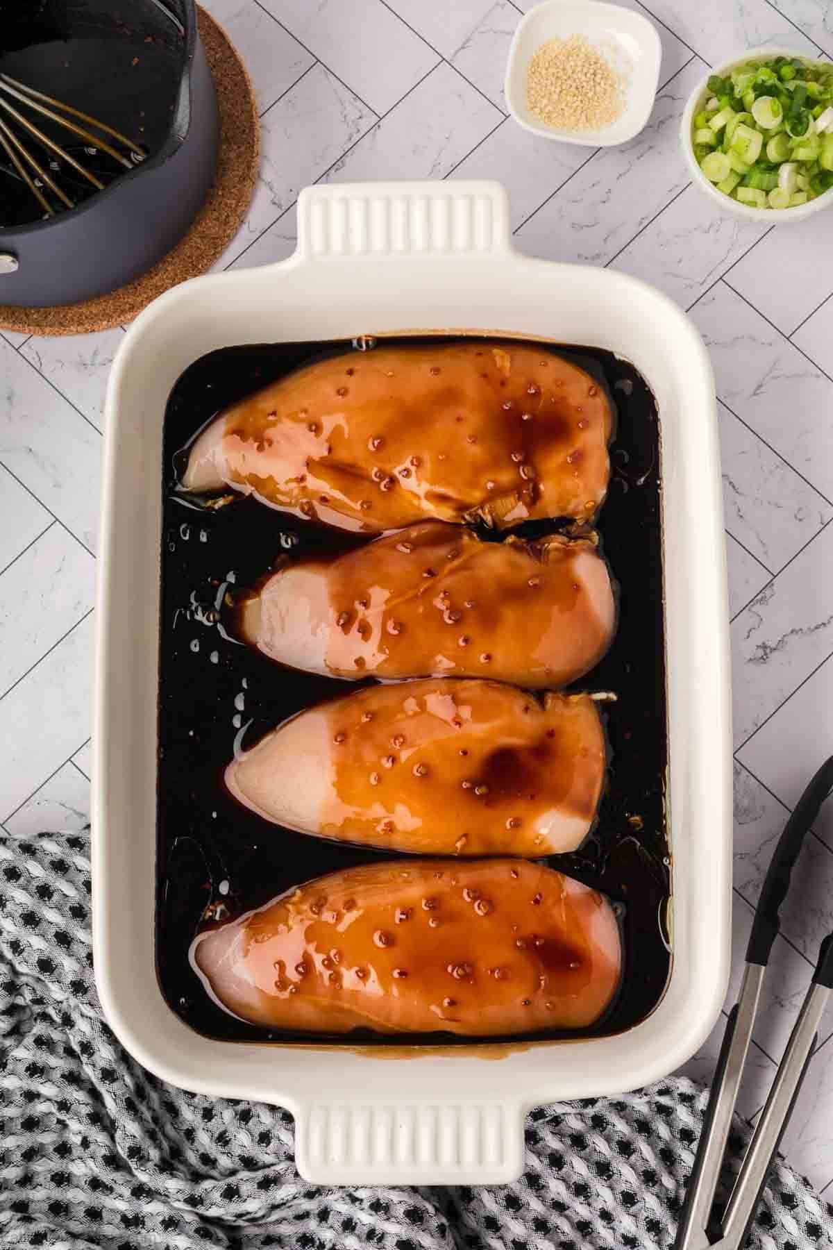 Four raw chicken breasts coated in a brown marinade, ready for a baked teriyaki chicken recipe, sit in a white rectangular baking dish. Surrounding items include a checkered towel, metal tongs, green onions, sesame seeds, and a saucepan with a whisk.