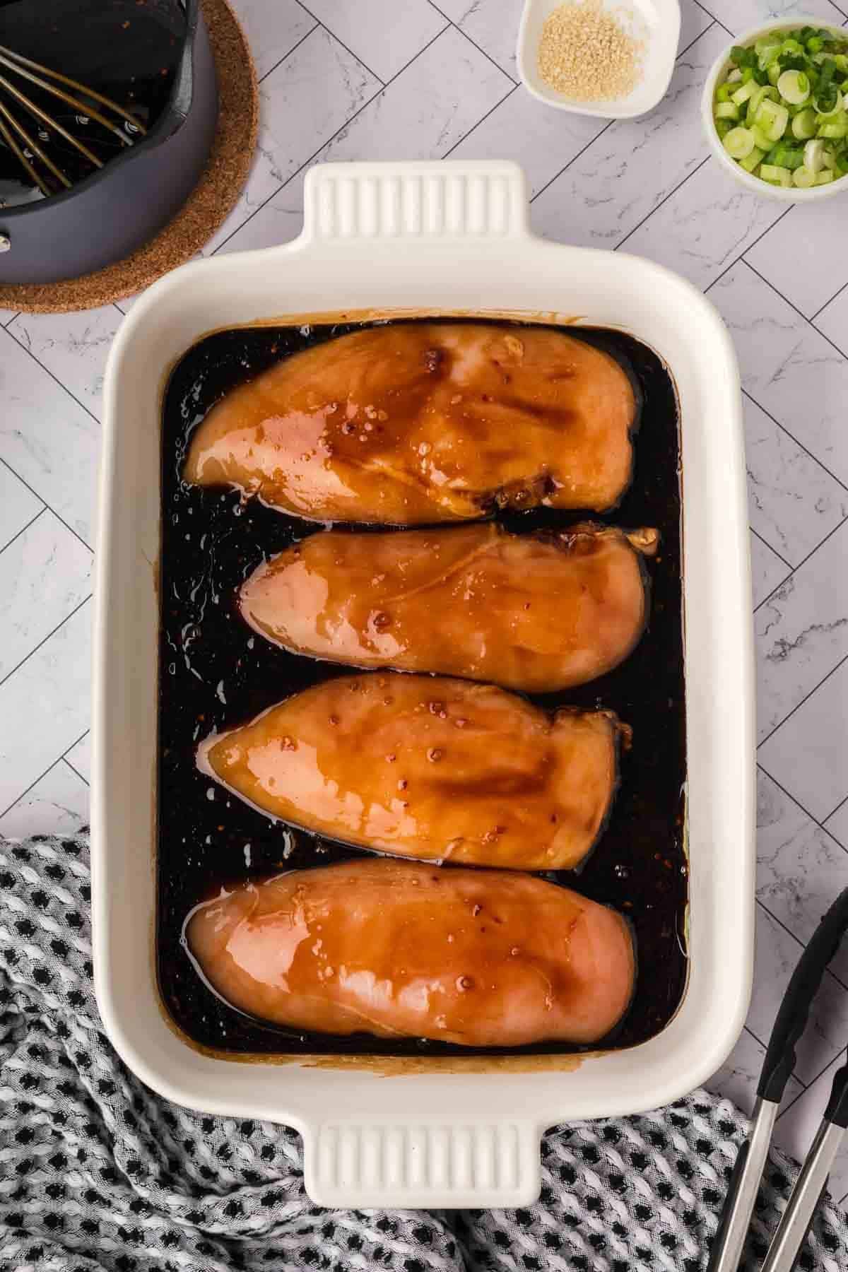 Four raw chicken breasts coated in a savory baked teriyaki chicken recipe marinade sit in a white rectangular baking dish. The dish rests on a checkered cloth, with tongs, sliced green onions, and sesame seeds nearby on the tiled countertop.
