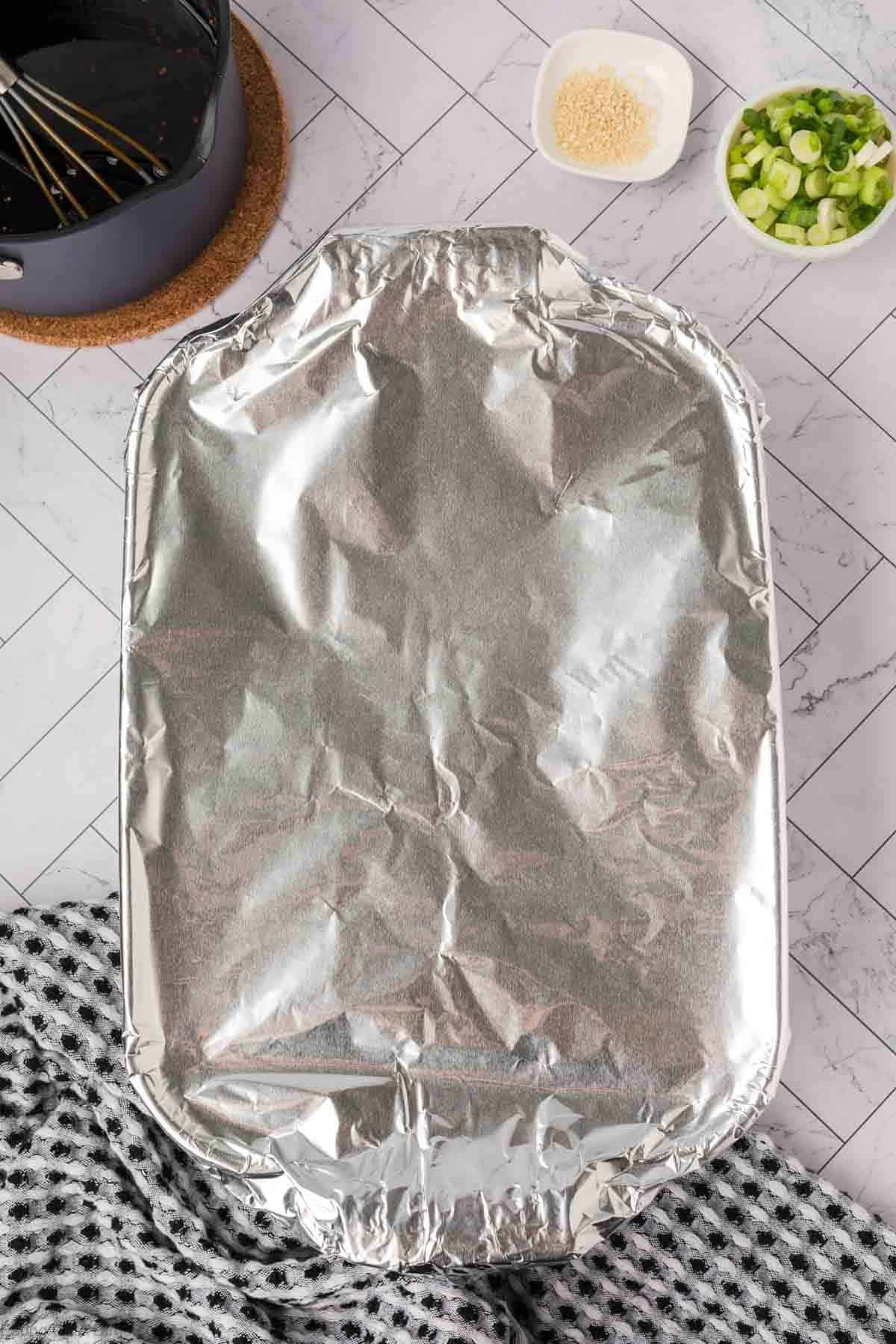 A rectangular baking dish covered with aluminum foil sits on a tiled surface, ready for a delicious baked teriyaki chicken recipe. Nearby are a pot of spaghetti, chopped green onions, grated cheese, and a patterned cloth in the foreground.