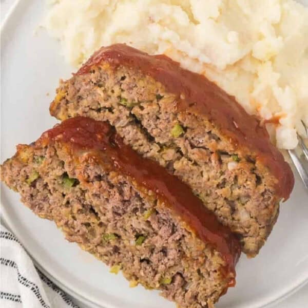 Two slices of Cracker Barrel Meatloaf Recipe topped with ketchup glaze sit on a plate next to a serving of mashed potatoes, with a fork on the side.