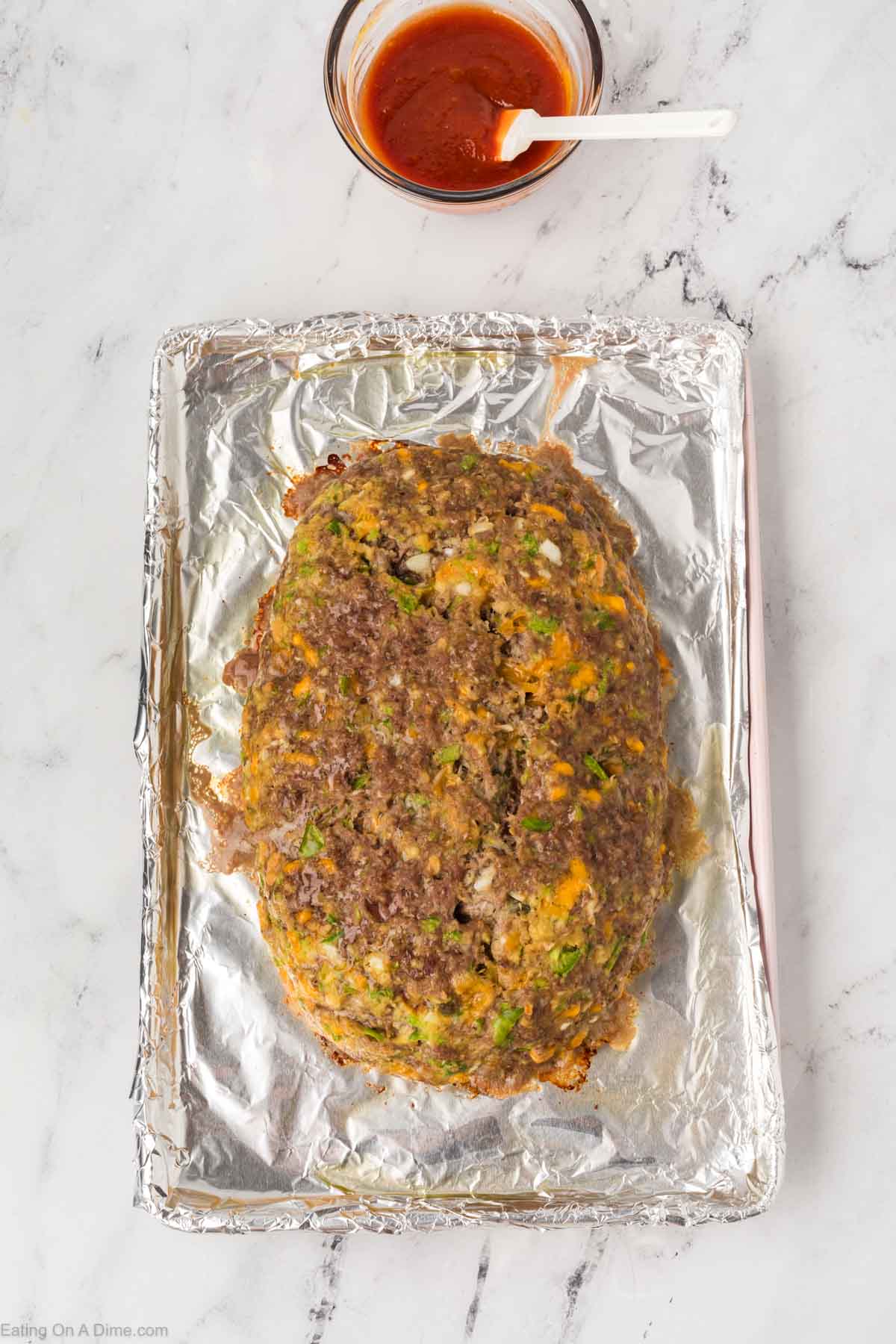 A cooked meatloaf, inspired by the Cracker Barrel Meatloaf Recipe, sits on a foil-lined baking sheet with a small bowl of red sauce and a white spoon next to it on a white marble surface.
