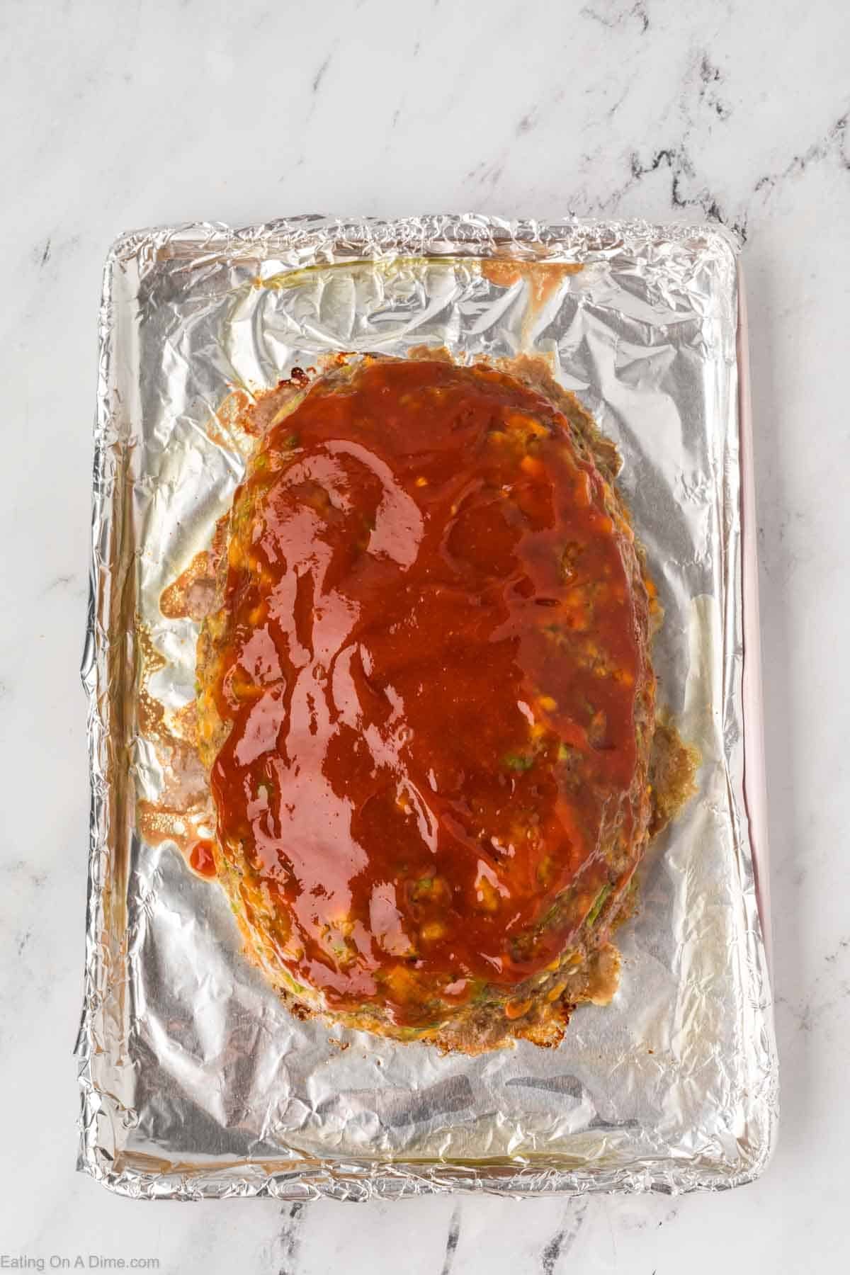 A baked meatloaf, inspired by the Cracker Barrel Meatloaf Recipe, sits covered in a shiny red glaze on a foil-lined baking sheet atop a marble countertop.