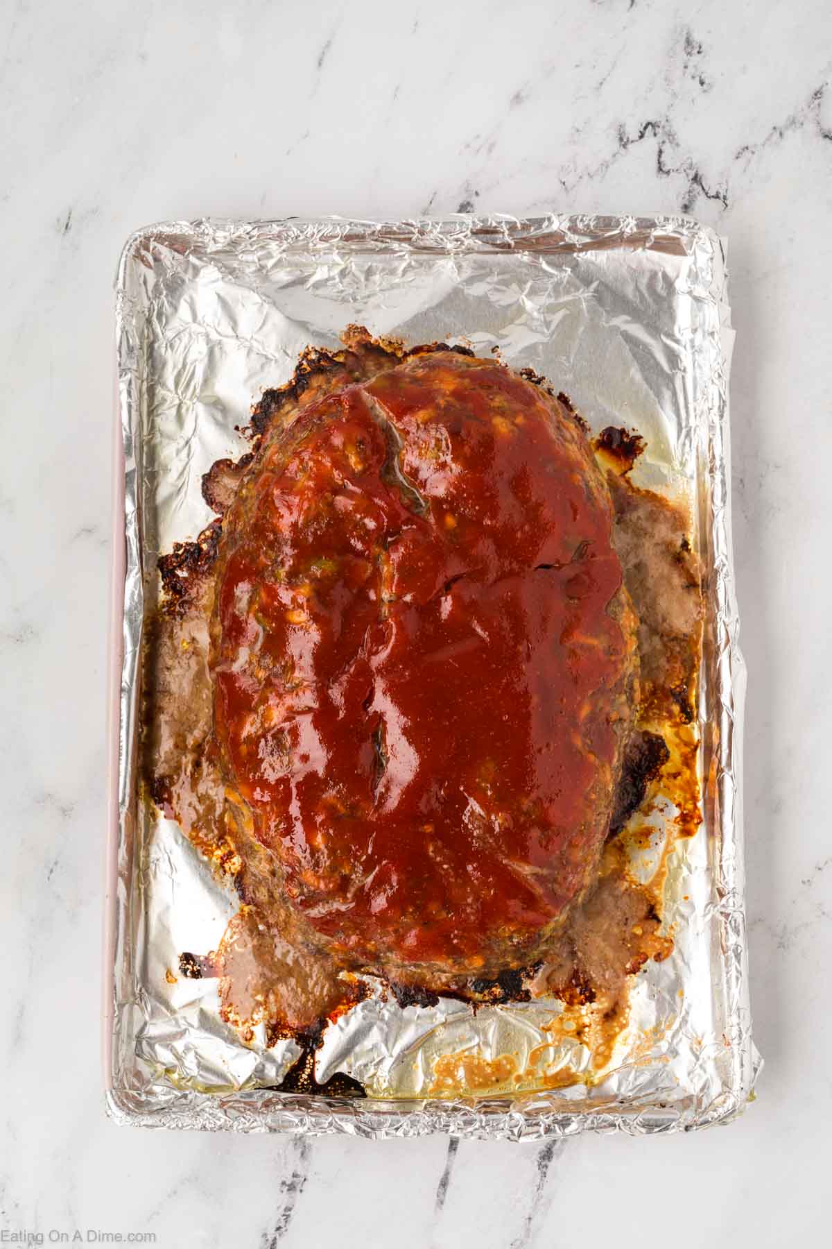 A baked meatloaf, inspired by the Cracker Barrel Meatloaf Recipe, is covered in shiny red glaze and sits on a foil-lined baking sheet atop a marble surface.