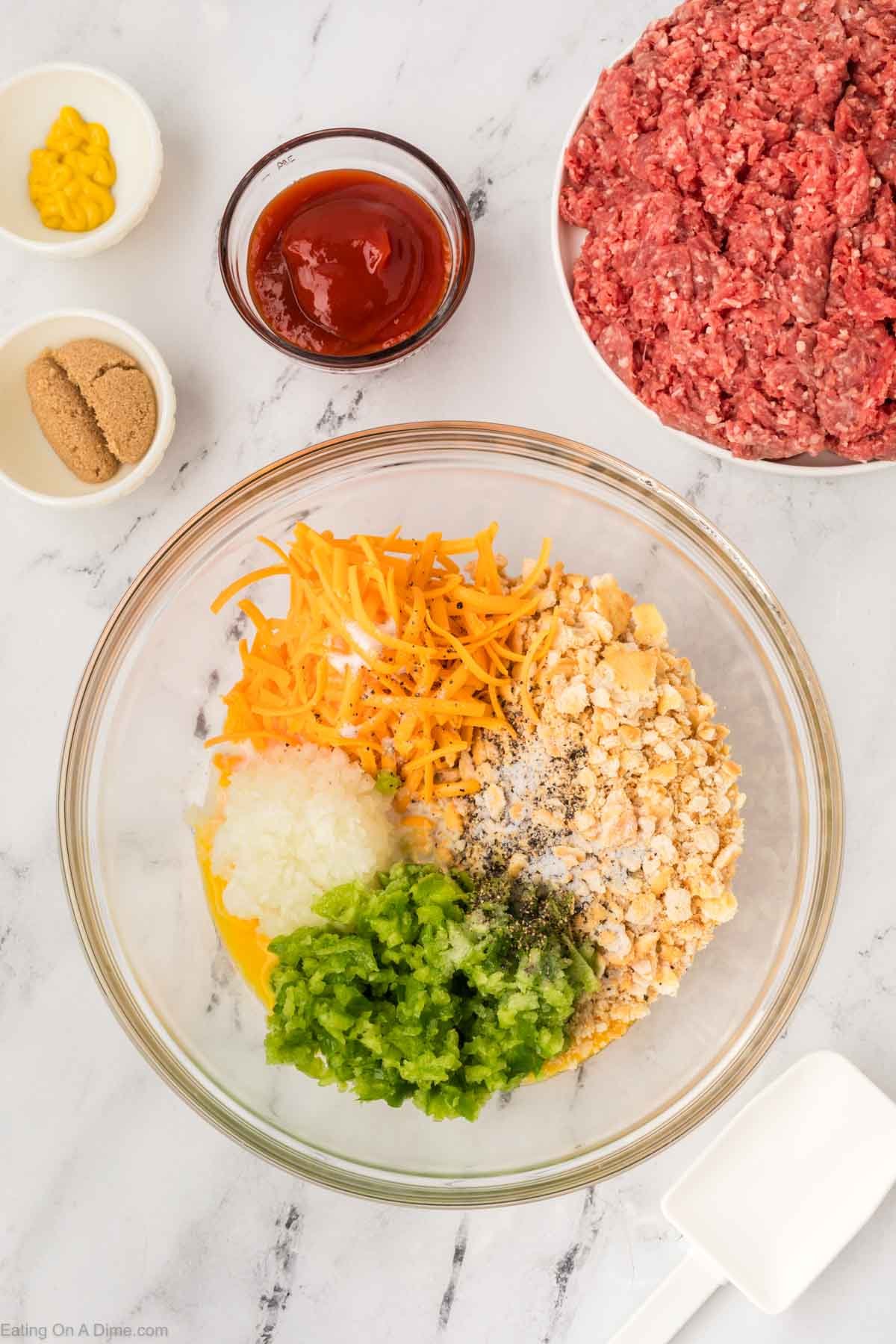 A glass bowl holds shredded cheddar, crushed crackers, diced onion, chopped green pepper, black pepper, and salt—ingredients for a classic Cracker Barrel Meatloaf Recipe. Surrounding it are bowls of ground beef, ketchup, mustard, and brown sugar.