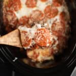 Close up image of pizza casserole in a crock pot with a serving on a wooden spoon.