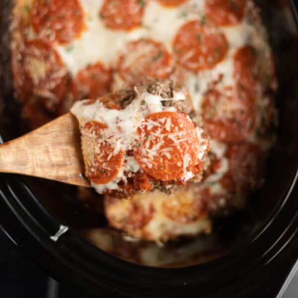 Close up image of pizza casserole in a crock pot with a serving on a wooden spoon.