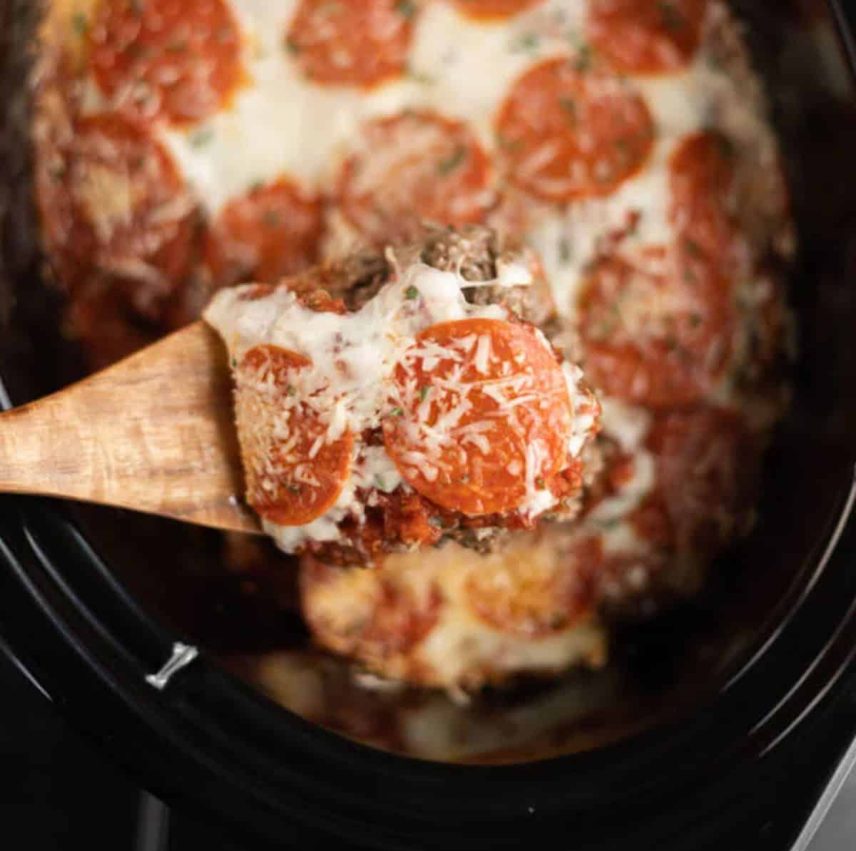 Close up image of pizza casserole in a crock pot with a serving on a wooden spoon.