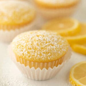 A low calorie lemon cupcake in a white wrapper, topped with powdered sugar, sits in focus with sliced lemons and two more cupcakes blurred in the background.