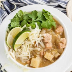 A bowl of Crock Pot Green Chili Stew with pork, potatoes, shredded cheese, lime wedges, and fresh cilantro sits on a striped towel.