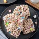 Three Lucky Charms Treats made with colorful cereal pieces and mini marshmallows are stacked on a black plate, with a few marshmallows scattered nearby.