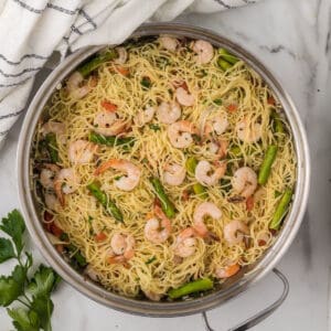 A pan filled with cooked angel hair pasta, shrimp, and green asparagus tips echoes the essence of an Olive Garden shrimp scampi recipe. The pasta is lightly tossed with herbs and small tomato pieces. A white striped towel and fresh parsley adorn the marble surface beside the pan.