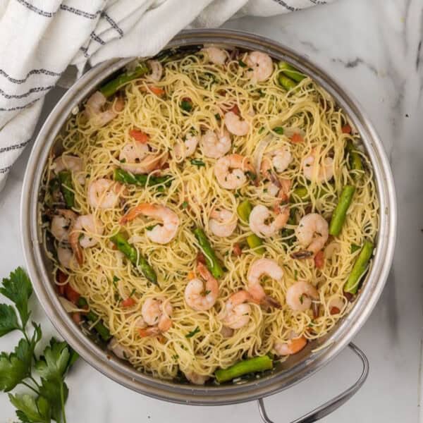 A pan filled with cooked angel hair pasta, shrimp, and green asparagus tips echoes the essence of an Olive Garden shrimp scampi recipe. The pasta is lightly tossed with herbs and small tomato pieces. A white striped towel and fresh parsley adorn the marble surface beside the pan.