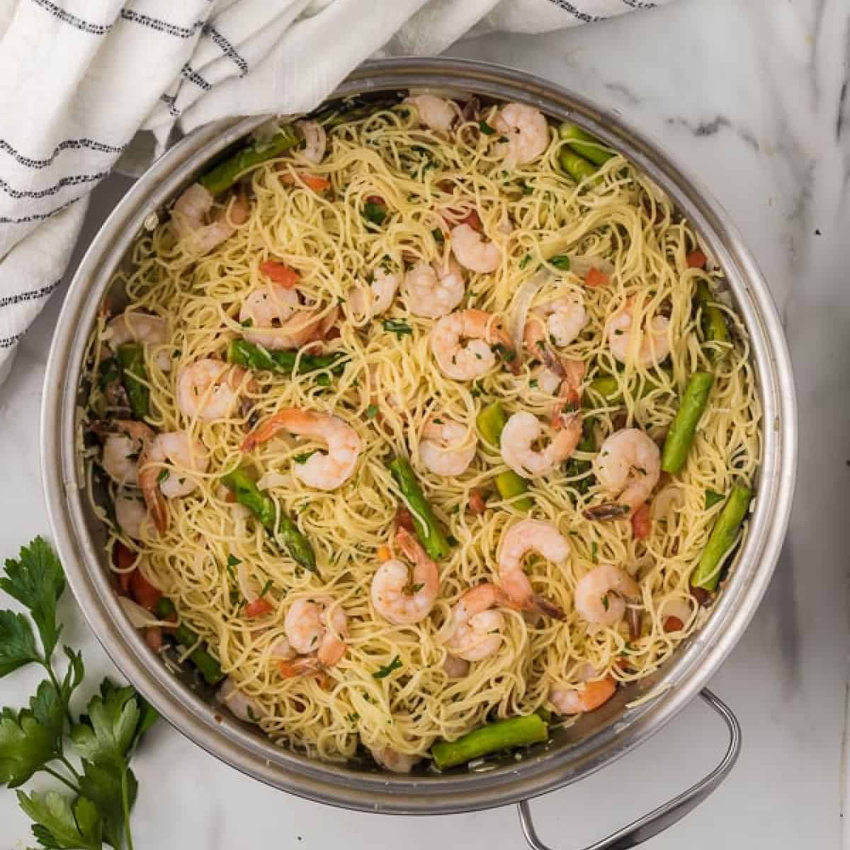 A pan filled with cooked angel hair pasta, shrimp, and green asparagus tips echoes the essence of an Olive Garden shrimp scampi recipe. The pasta is lightly tossed with herbs and small tomato pieces. A white striped towel and fresh parsley adorn the marble surface beside the pan.