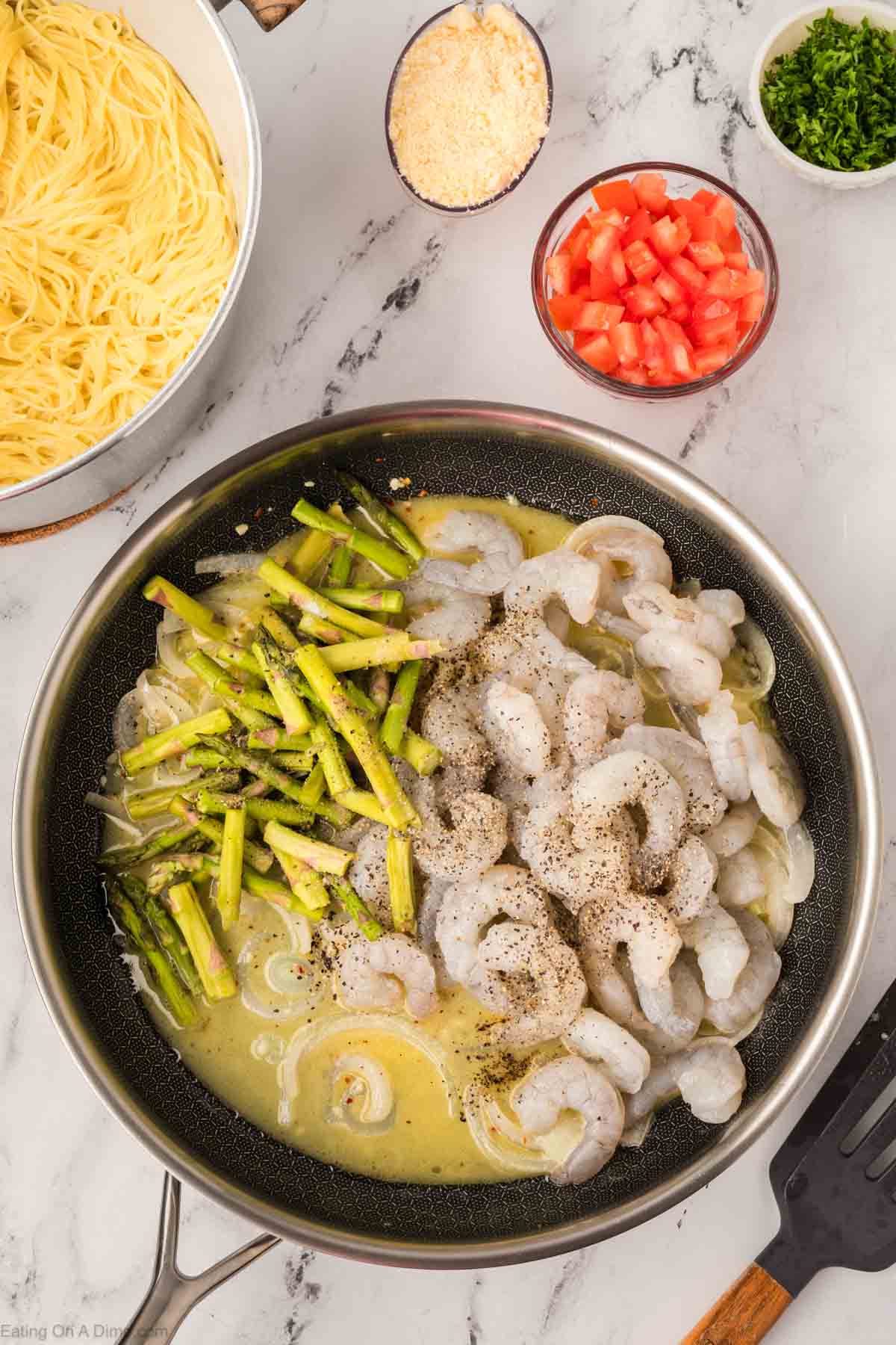A skillet with uncooked shrimp, asparagus, and seasonings in a creamy sauce inspired by an Olive Garden shrimp scampi recipe. Nearby are bowls of grated cheese, chopped tomatoes, fresh herbs, and a pot of cooked pasta on a marble countertop.