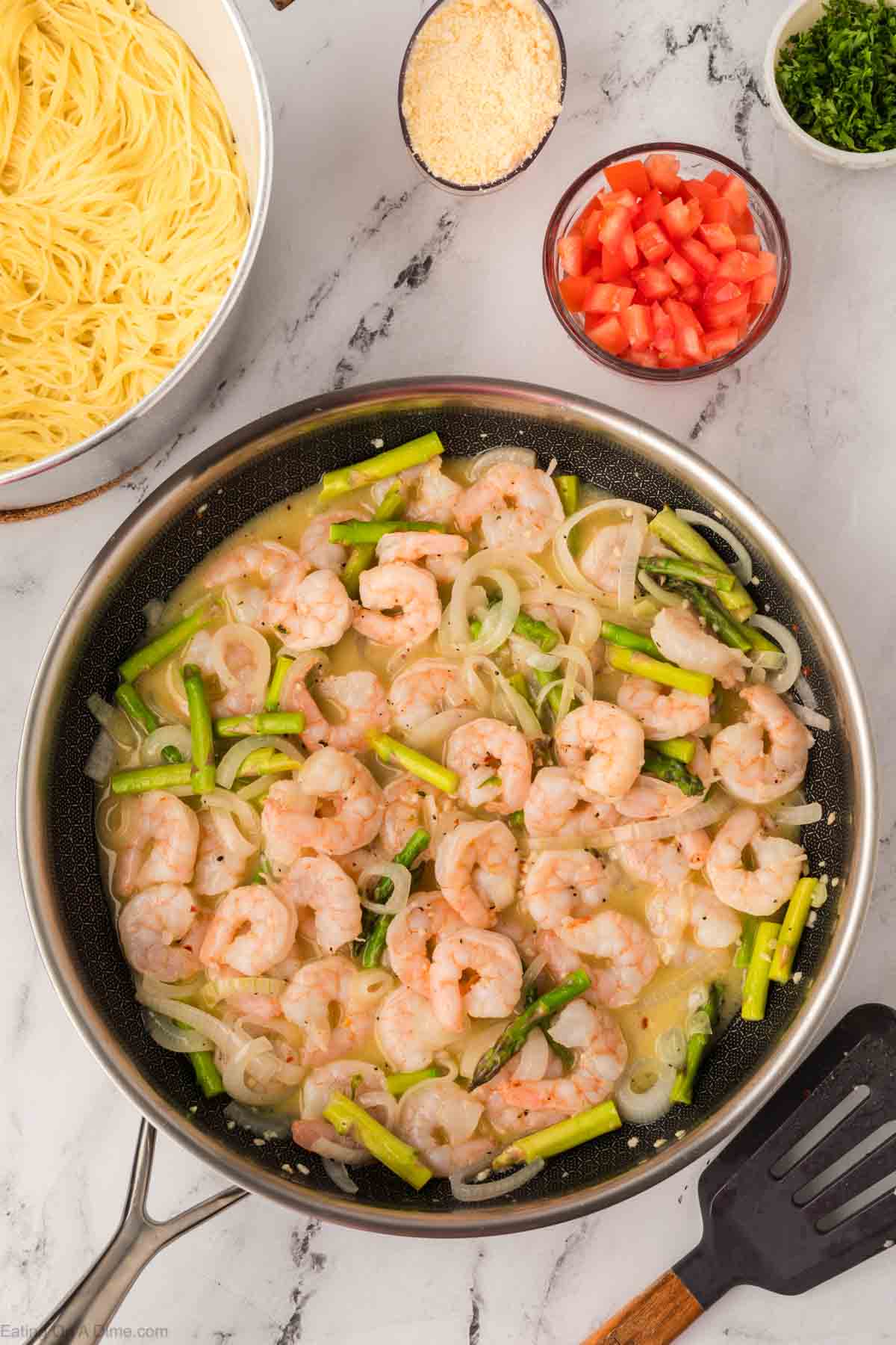 A skillet with shrimp, asparagus, and onions simmering in a creamy sauce sits on a marble countertop, reminiscent of an Olive Garden shrimp scampi recipe. Nearby are bowls of cooked pasta, diced tomatoes, grated cheese, and chopped herbs.