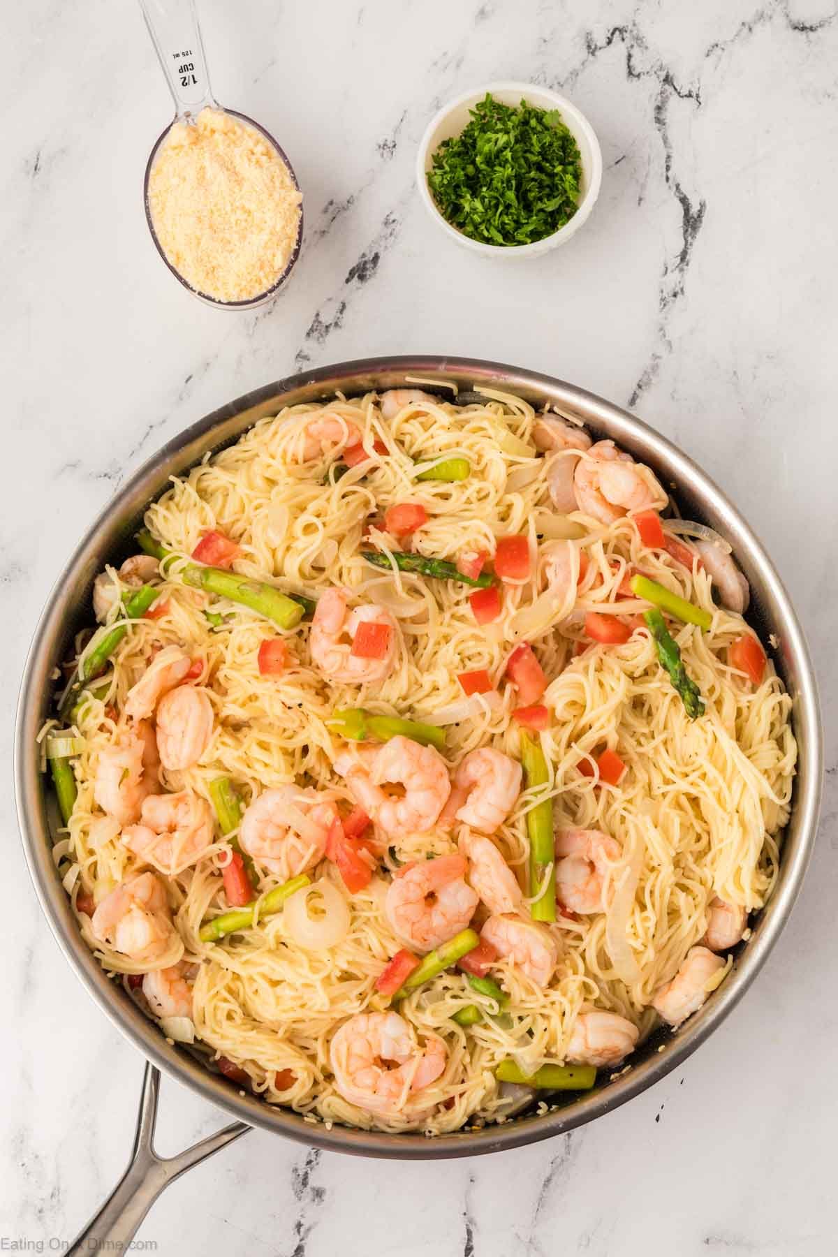 A skillet filled with shrimp scampi pasta, inspired by the Olive Garden shrimp scampi recipe, is mixed with asparagus and tomatoes. Nearby sit a small bowl of chopped parsley and a measuring cup of grated Parmesan cheese.