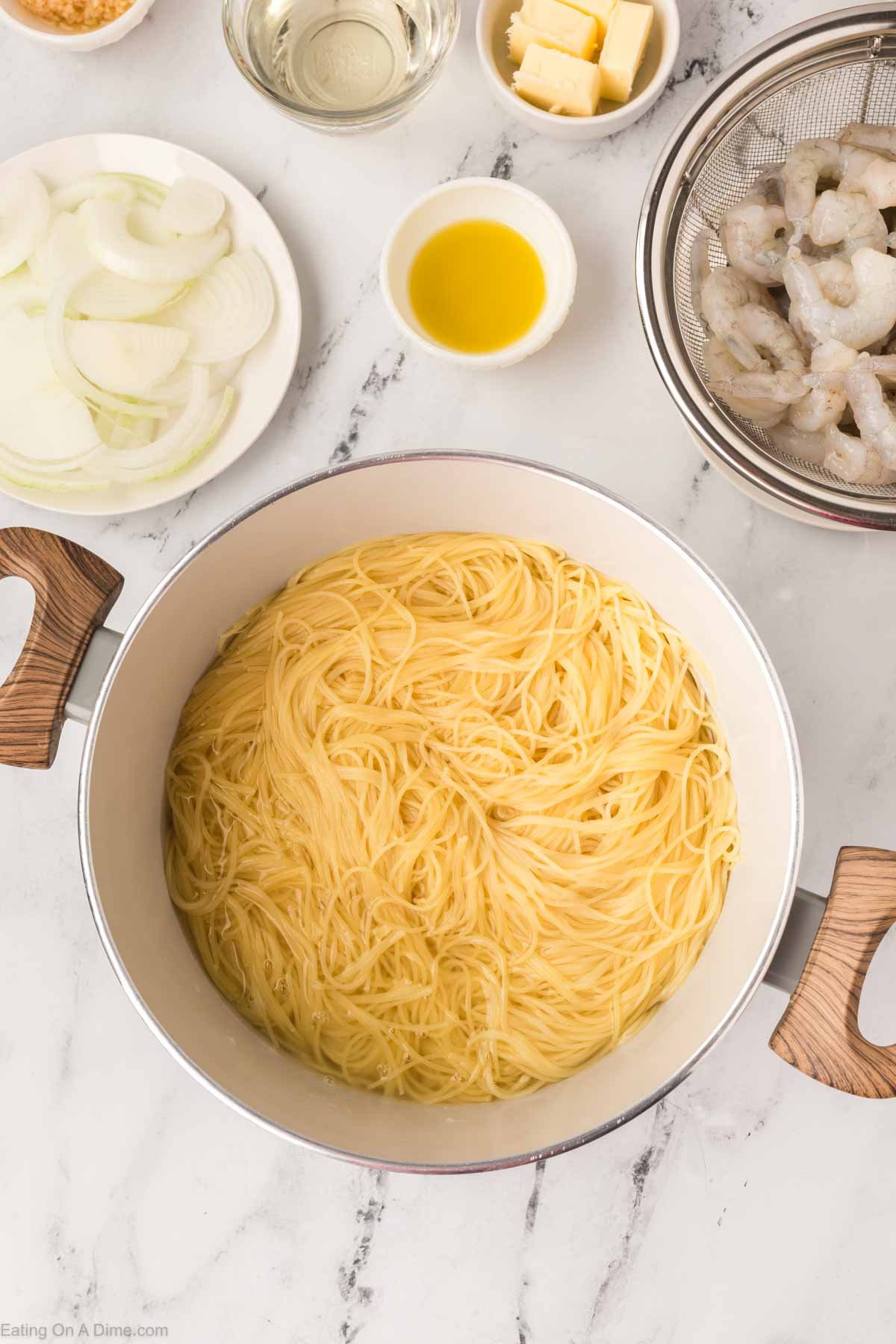 A white pot filled with cooked spaghetti sits on a marble counter, surrounded by bowls of sliced onions, raw shrimp, olive oil, butter, and a dish of minced garlic—perfect for recreating the Olive Garden shrimp scampi recipe at home.