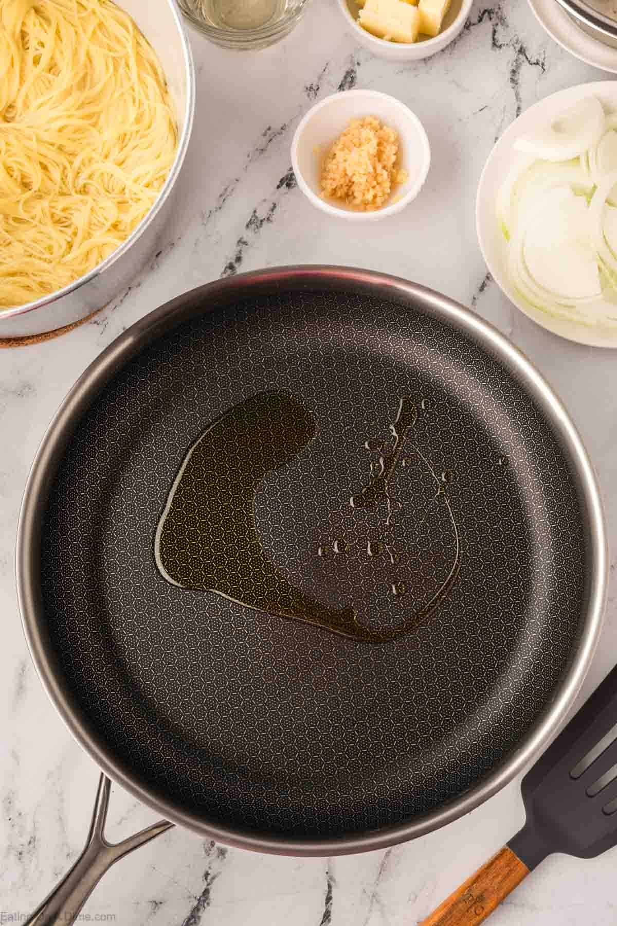 A frying pan with a drizzle of oil sits on a marble countertop, surrounded by bowls of uncooked pasta, sliced onions, minced garlic, and cubed butter—just the setup for an Olive Garden shrimp scampi recipe. A spatula rests nearby.