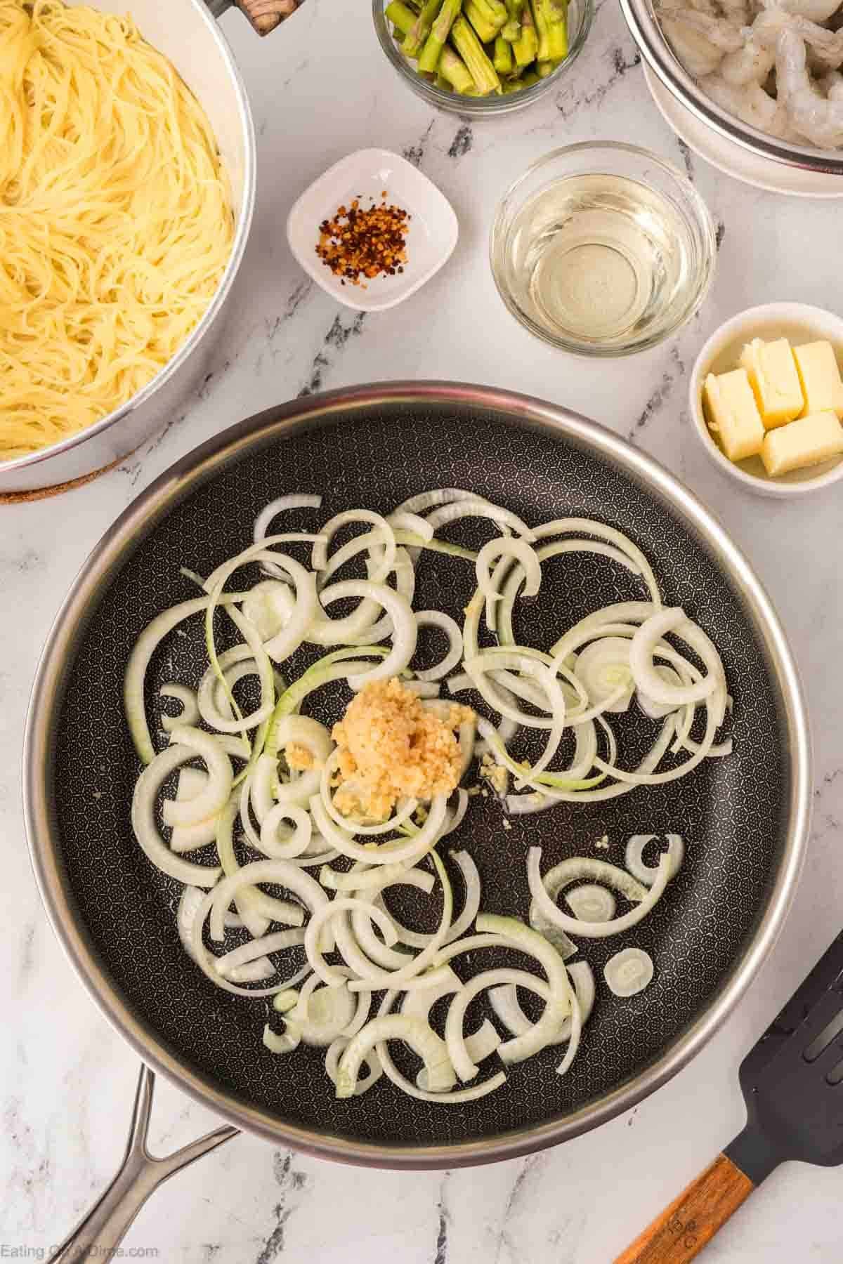 A frying pan with sliced onions and minced garlic sits on a marble countertop, surrounded by bowls of cooked pasta, shrimp, butter, red pepper flakes, white wine, and cut asparagus—perfect for an Olive Garden shrimp scampi recipe.