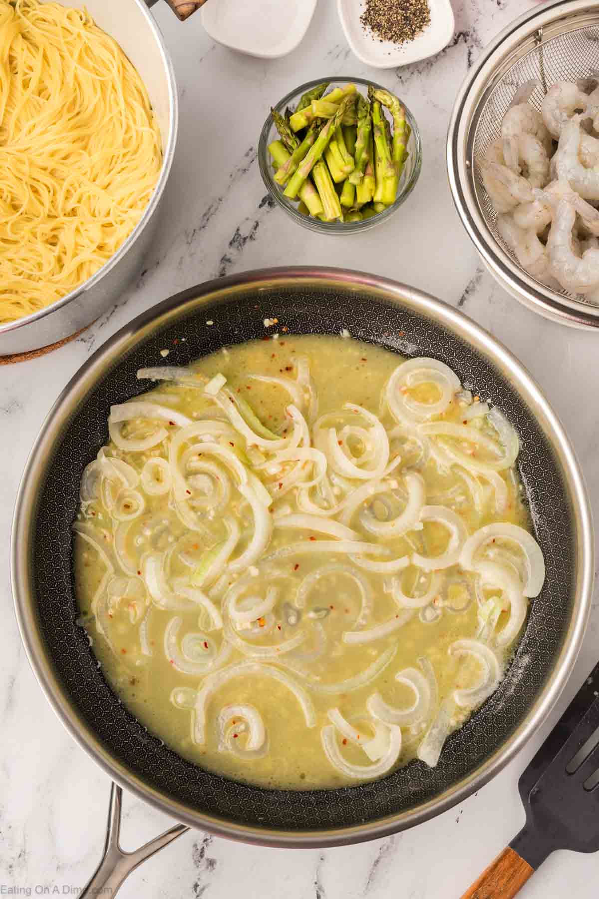 A skillet with sliced onions sautéing in a light sauce, surrounded by bowls of cooked pasta, raw shrimp, sliced asparagus, and seasonings on a marble countertop—perfect for creating an Olive Garden shrimp scampi recipe at home.