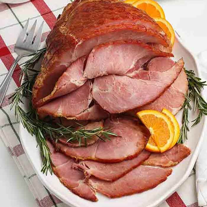A cooked spiral ham on a large white platter with diced oranges or fresh herbs on it 