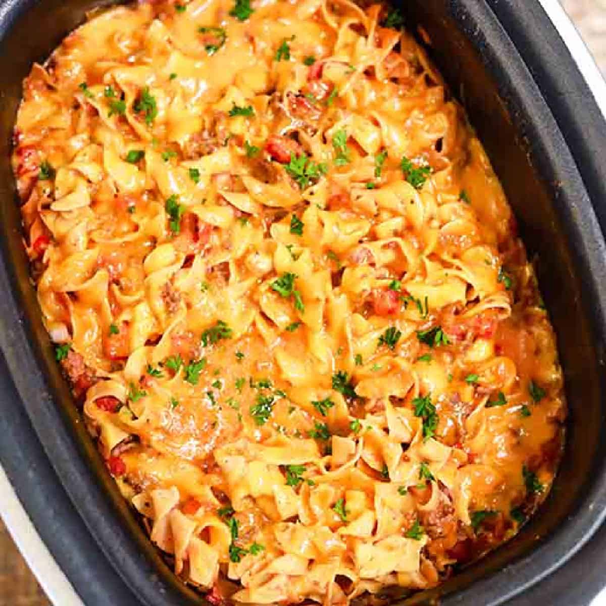 A close-up of a cheesy beef noodle casserole simmering in a crock pot. This savory dish features spiral noodles, melted cheese, and diced red tomatoes, all harmoniously mingling with chopped green herbs sprinkled on top.