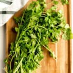 A bunch of fresh cilantro with green leaves and stems on a wooden cutting board, next to a white cloth with a black plaid pattern—perfect for anyone exploring the best cilantro substitutes in their recipes.