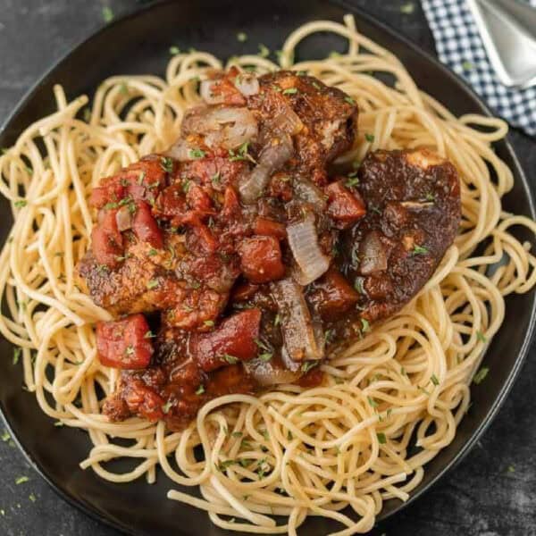 A black plate of cooked spaghetti topped with Slow Cooker Balsamic Chicken, diced tomatoes, onions, and herbs in a rich sauce, placed on a dark surface with a checkered napkin and utensils nearby.