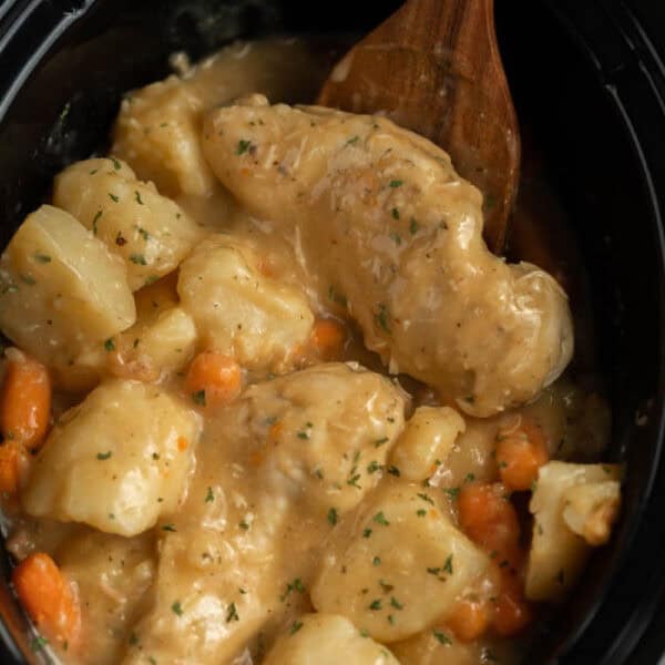 Crockpot Creamy Ranch Chicken, potatoes, and carrots simmer in a creamy sauce. A wooden spoon is partially submerged in the hearty stew, garnished with herbs.