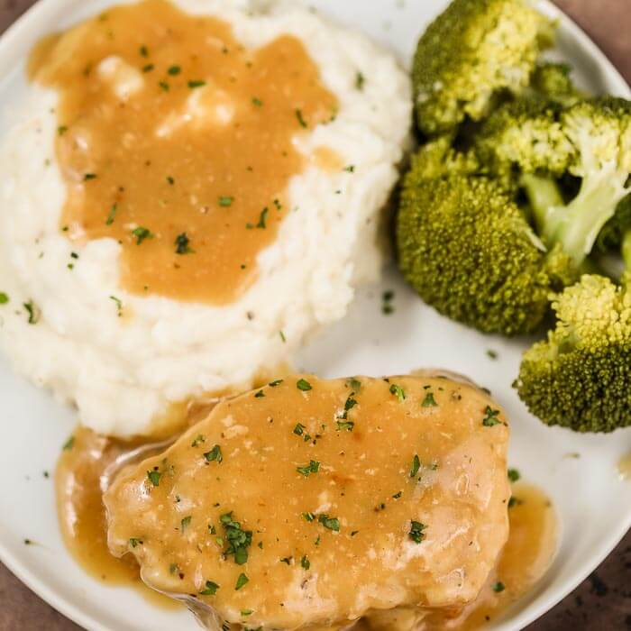 Close up image of pork chops and gravy with a side of mashed potatoes and broccoli on a white plate. 