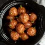 A close-up of Crock Pot Sweet and Spicy Meatballs glazed to perfection and garnished with chopped green onions, served in a black slow cooker.