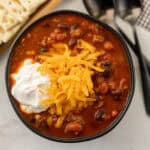 A bowl of crock pot vegetable and beef chili recipe topped with shredded cheddar cheese and a dollop of sour cream, with black and kidney beans visible. Crackers are on the side, and two spoons are placed nearby.