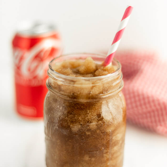 Close up image of a coke slushie in a mason jar with a straw and one can of coca-cola in the background