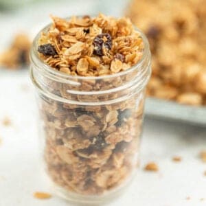 A glass jar filled with an easy homemade granola recipe, featuring oats, nuts, and raisins, sits on a light surface. Blurred granola on a tray is visible in the background.