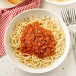 A bowl of spaghetti topped with a rich homemade tomato sauce, garnished with herbs. The dish rests on a table with a red and white checkered cloth nearby, evoking the charm of a homemade spaghetti sauce recipe. A fork and spoon sit beside the inviting bowl.