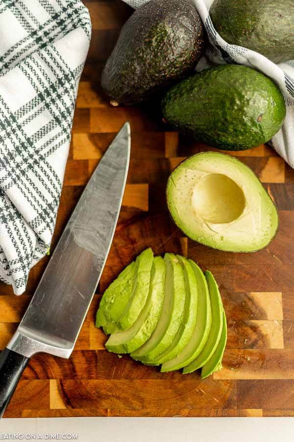 Close up image of sliced avocados with three whole avocados. Also a knife and a cutting board. 
