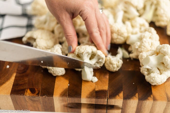 Cauliflower florets being cut on a cutting board with a knife.