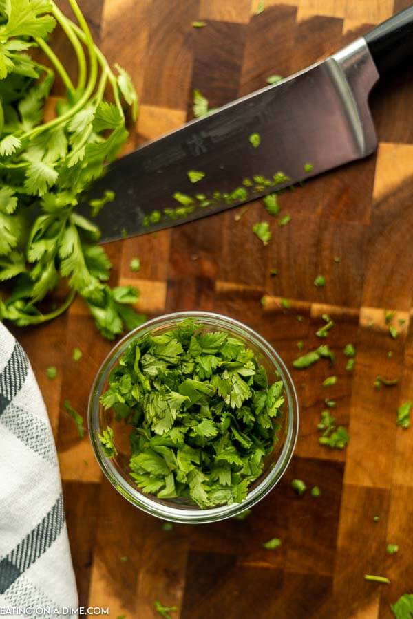 Chopped cilantro in a small bowl with cilantro leaves above it and a large knife laying next to it 