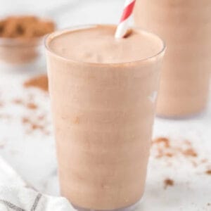 A glass filled with a creamy chocolate keto shake recipe, topped with a red and white striped straw. Cocoa powder is scattered in the background on a white surface.