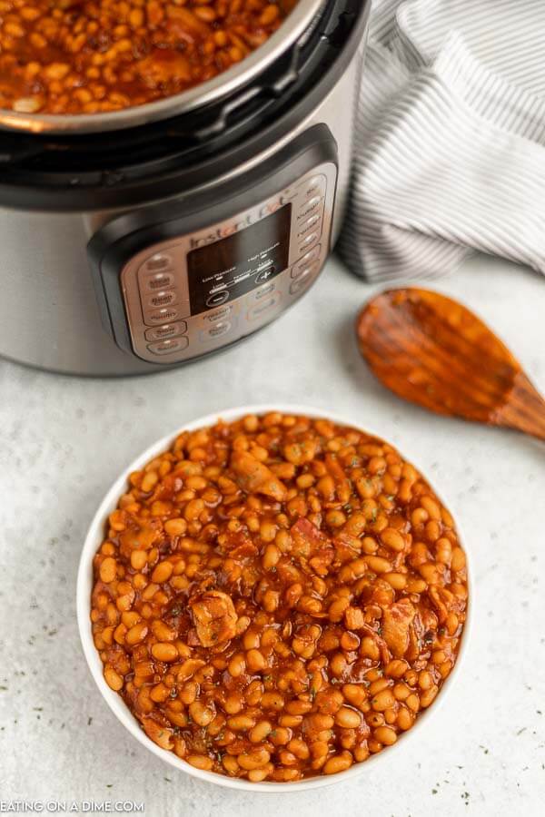 Close up image of baked beans in a white bowl with an instant pot and a wooden spoon. 