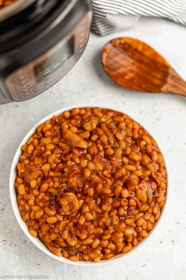 Close up image of baked beans in a white bowl with an instant pot and a wooden spoon. 